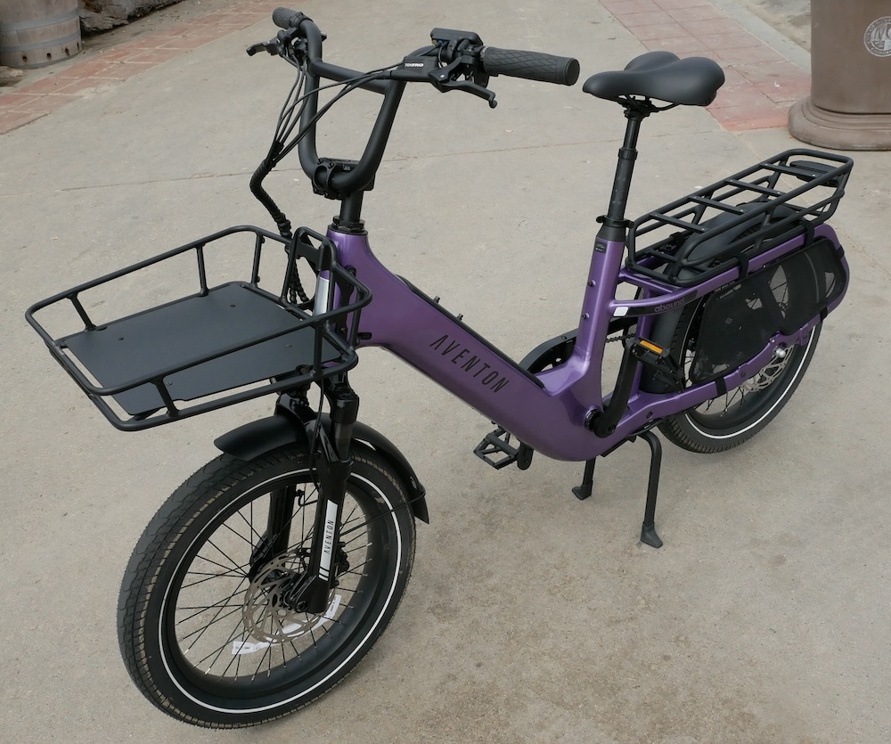 A light purple cargo bike with a front rack and rear rack, standing on a double kickstand. The bike says Aventon on the side.