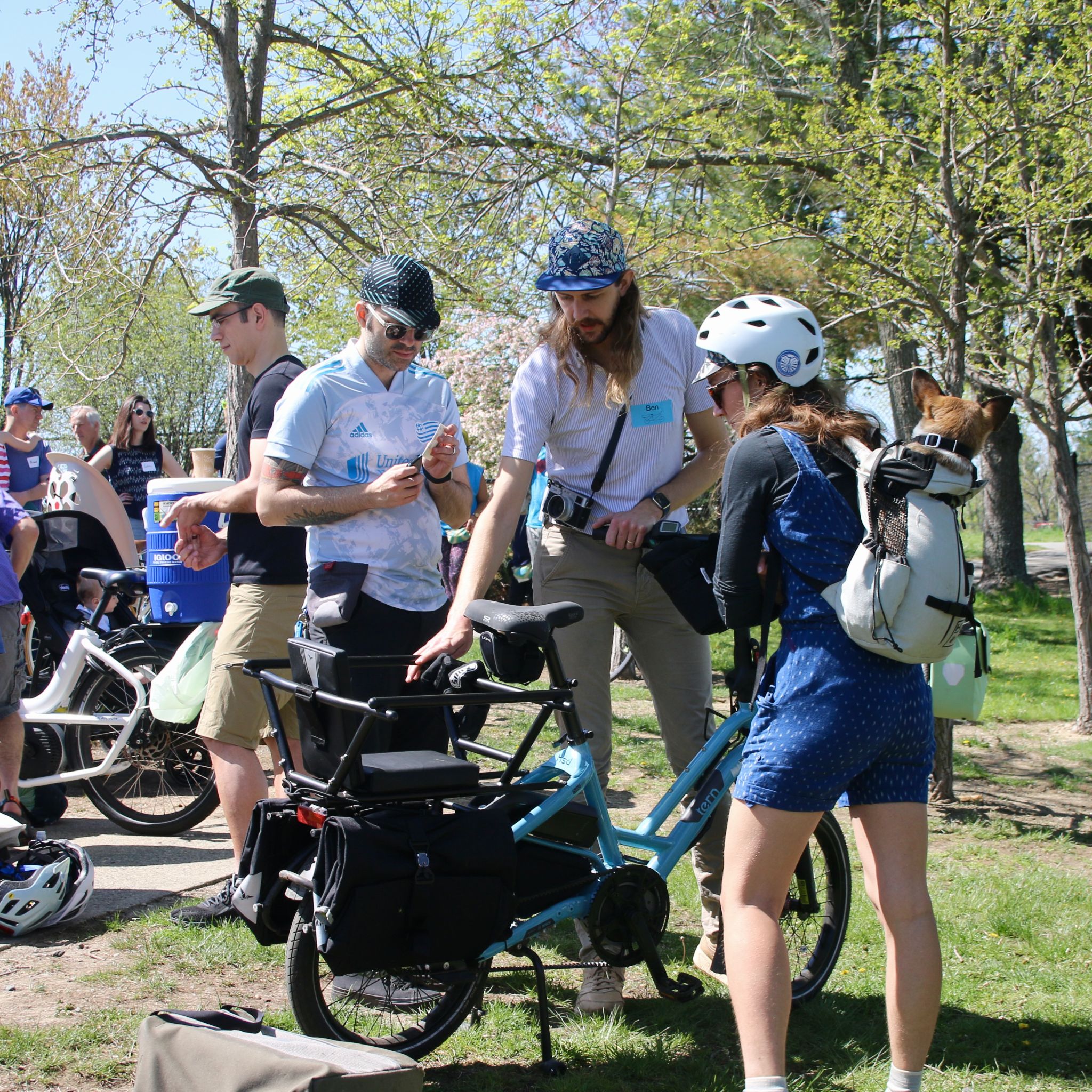 a group of people gathered around bicycles in a park setting on a sunny day. Some are engaged in conversation while checking out a blue cargo bike. One individual is holding a smartphone, and another has a dog peeking out from a backpack.